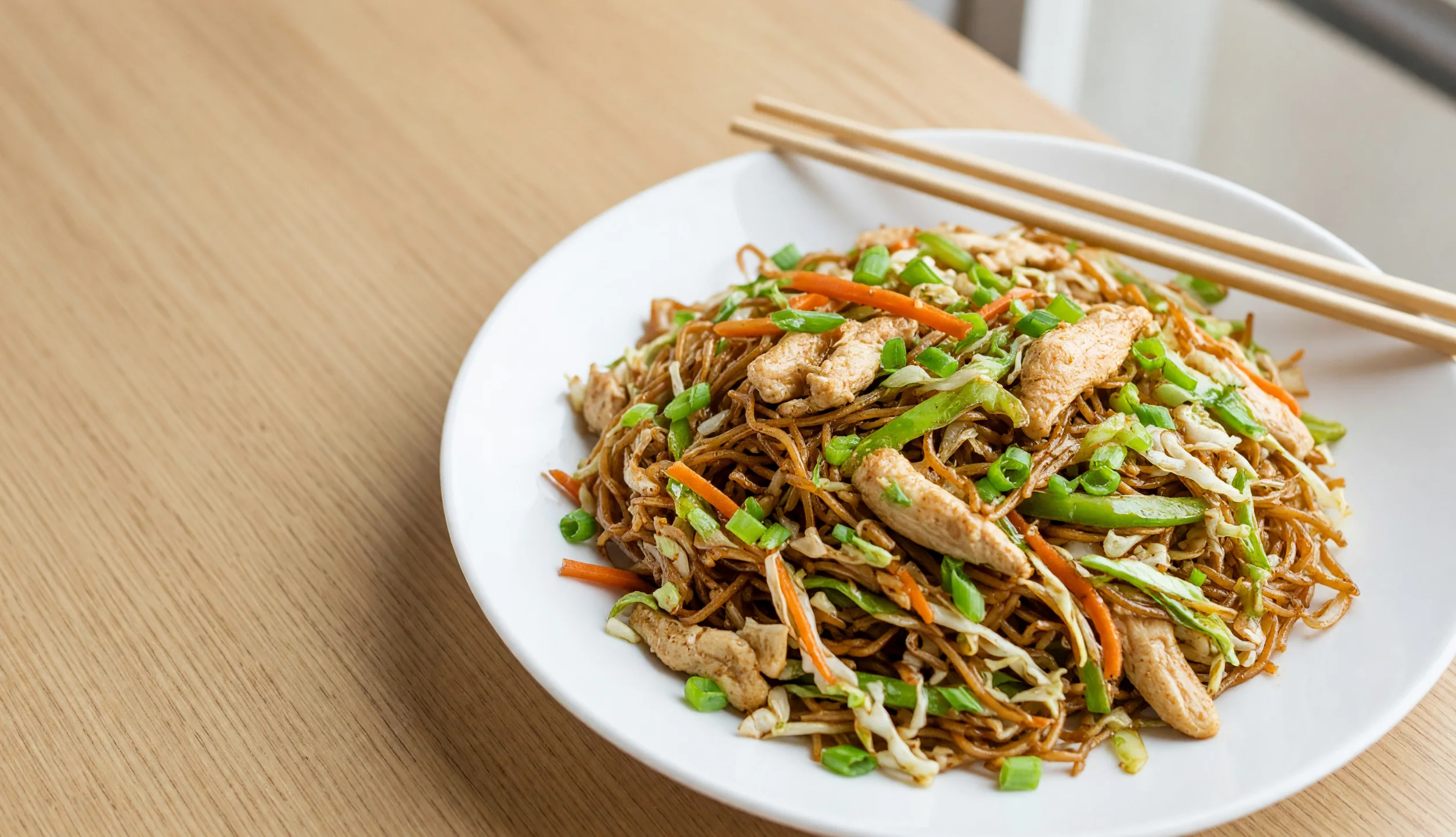 A plate of chicken Hakka noodles with vegetables and chopsticks on a light wood table