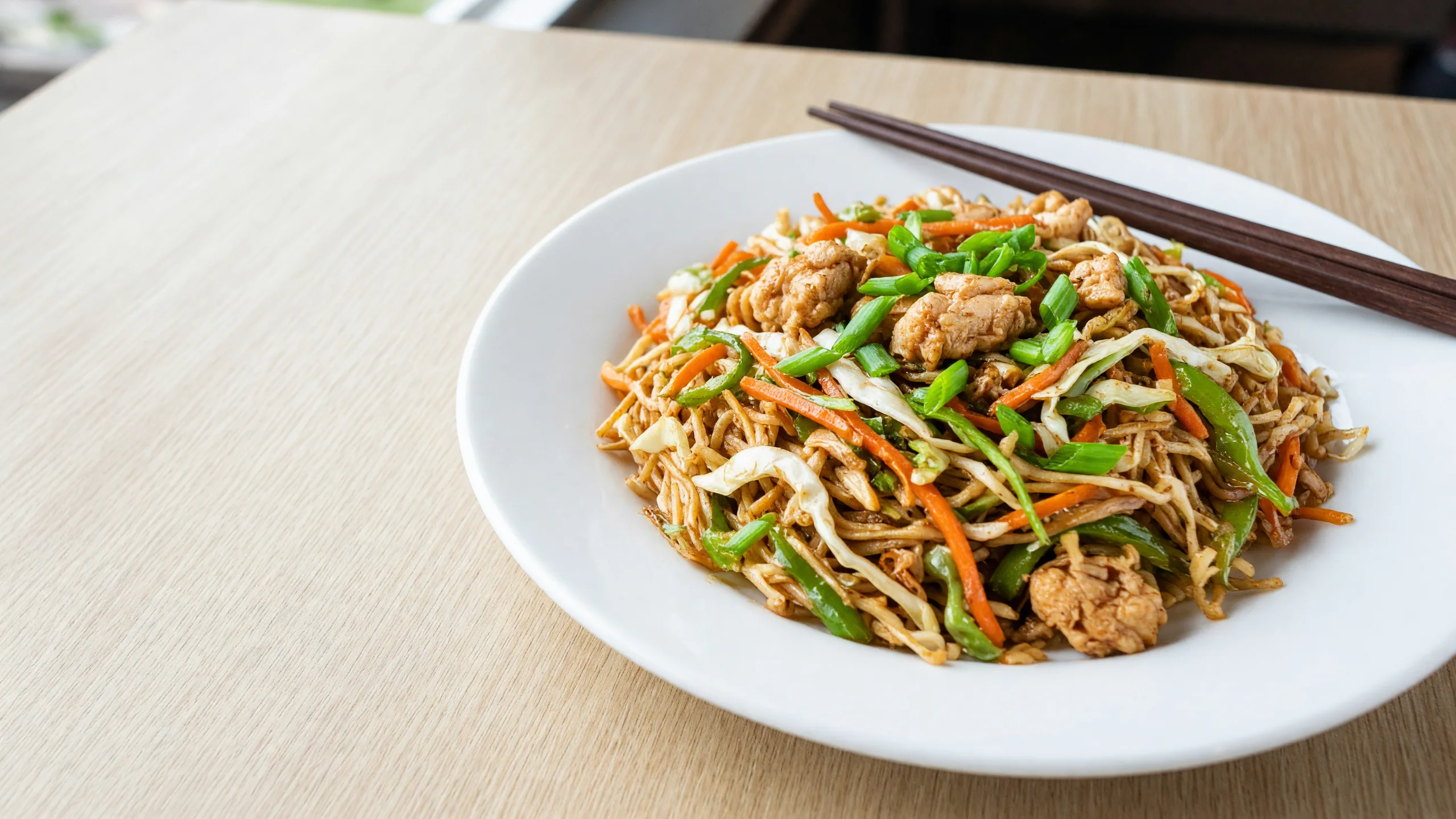 A white plate of chicken Hakka noodles with cabbage, carrot and green pepper, chopsticks on the plate